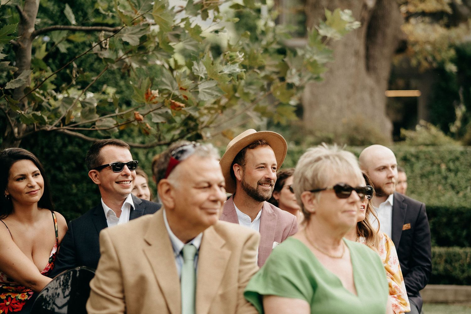 A group of wedding guests smile and laugh outdoors in a garden setting on a sunny afternoon.