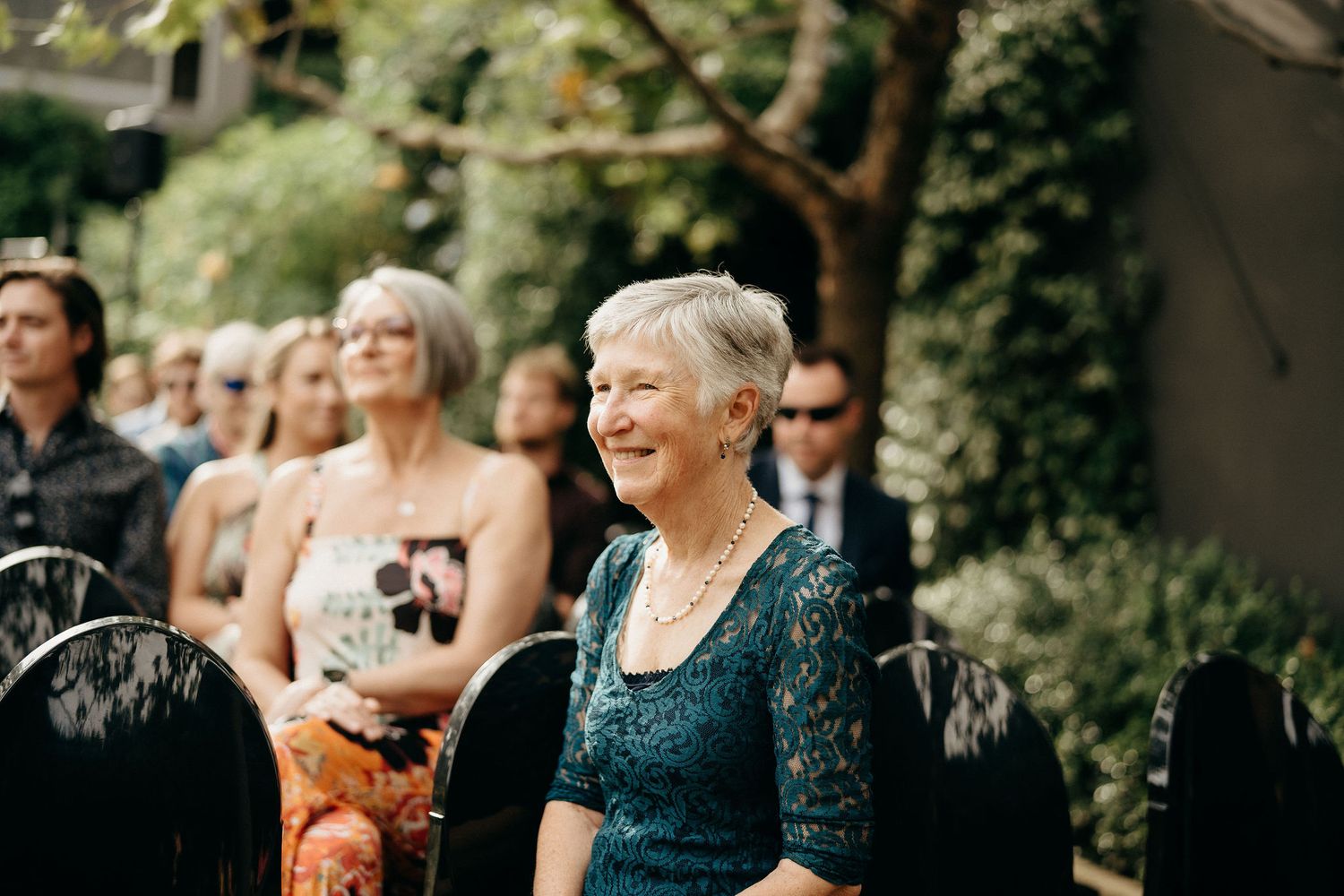 A guest in a navy lace dress watches the ceremony with joy at an outdoor wedding.