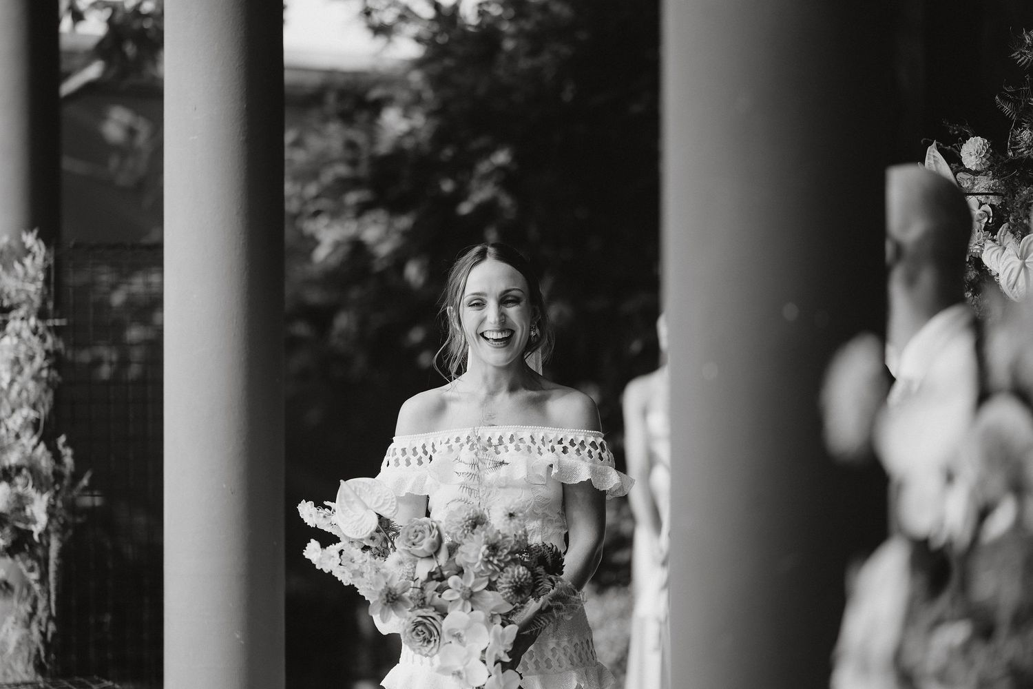 Black and white photo of a bride holding a floral bouquet while standing between elegant columns outdoors.