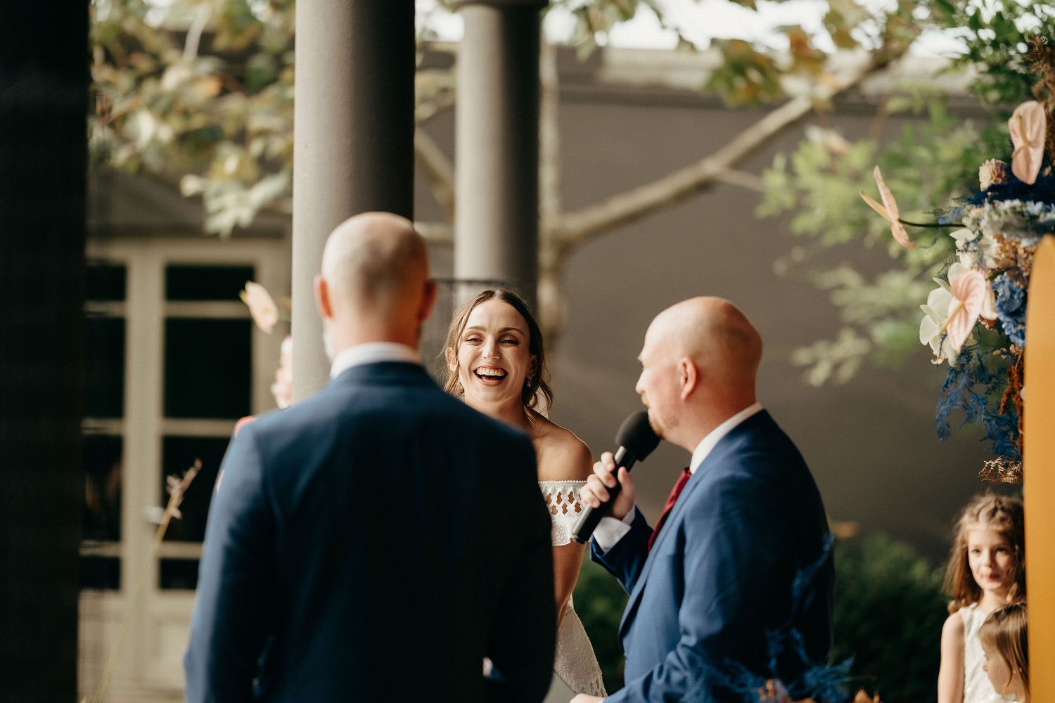 A smiling minister conducts an outdoor wedding ceremony under a covered porch area on a sunny day.