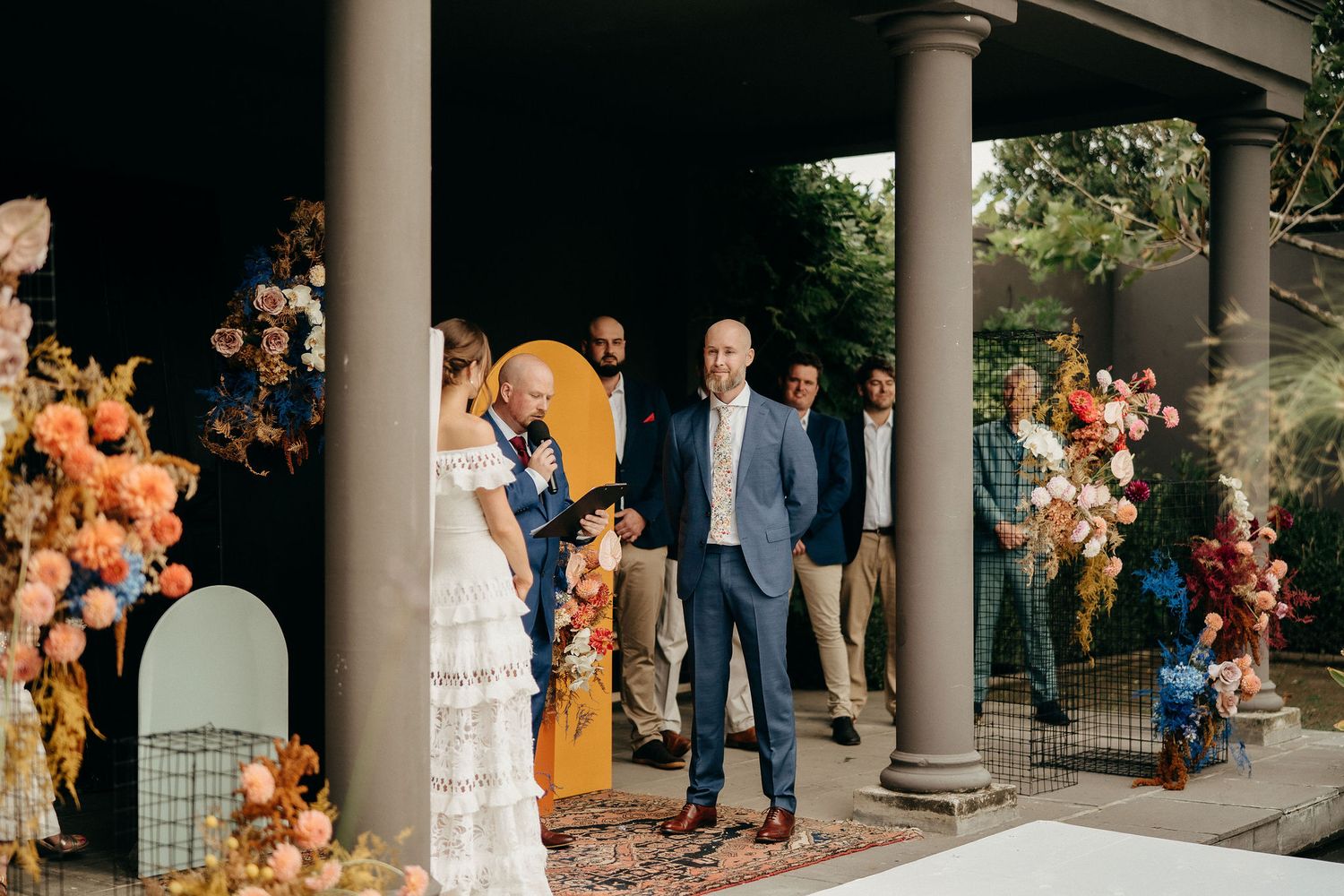 Elegant outdoor wedding ceremony with stone columns and vibrant orange floral arrangements adorning the entrance.