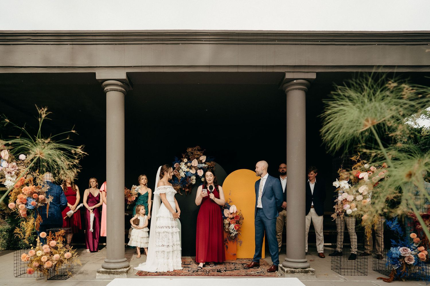 An outdoor wedding ceremony takes place under a columned portico adorned with elegant floral arrangements and a gold arch.