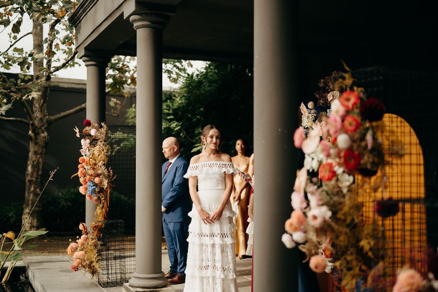 An outdoor wedding ceremony under a columned portico decorated with autumn-colored floral arrangements and balloons.