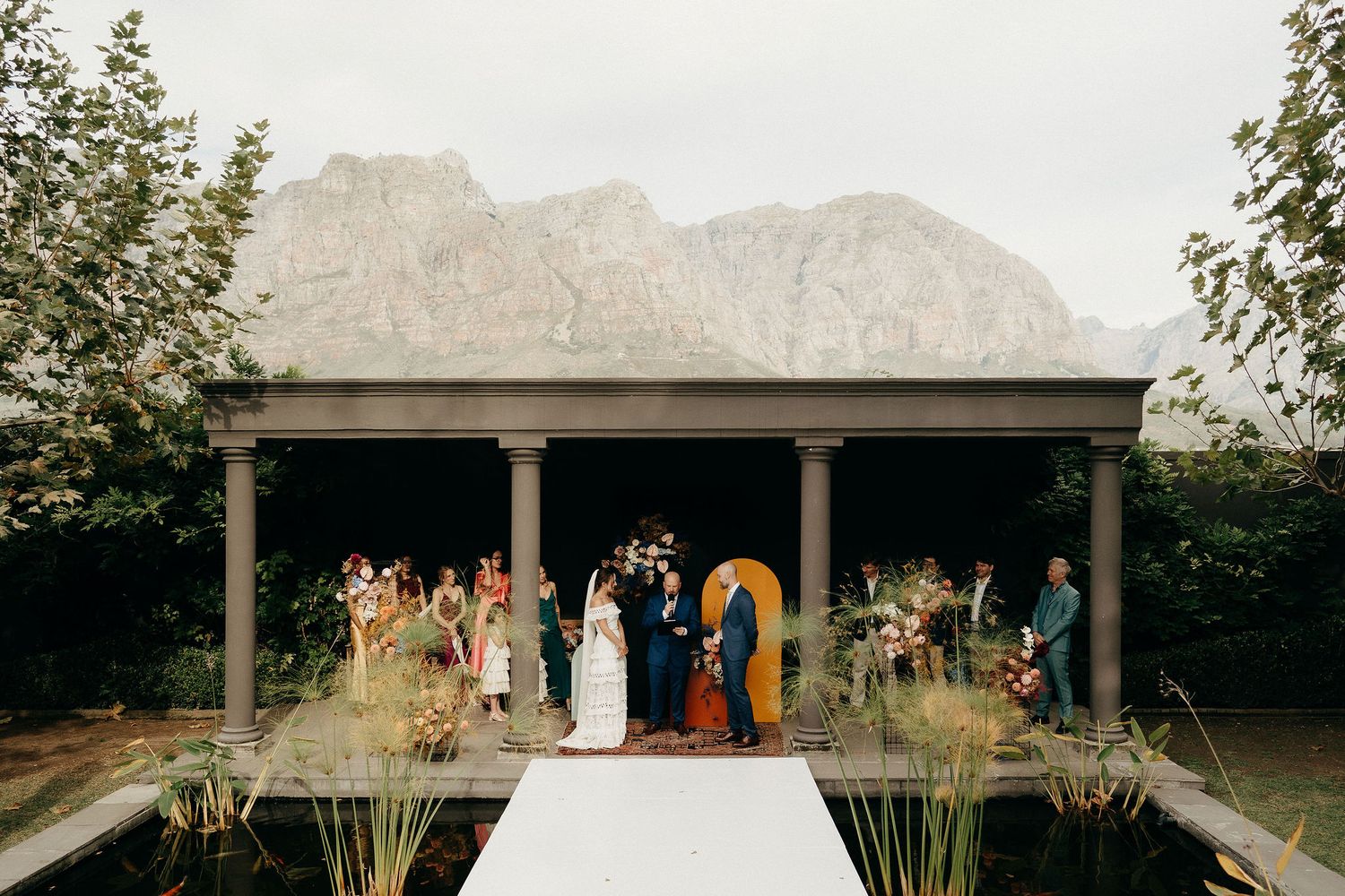 An outdoor wedding ceremony takes place under a covered pavilion against dramatic mountain backdrop in a garden setting.