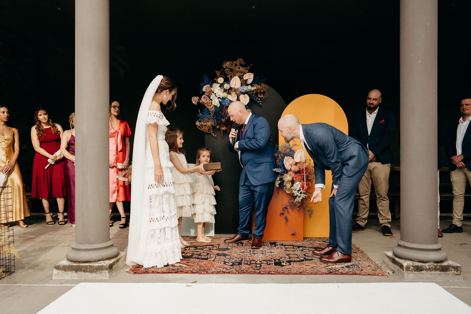 Two people stand at a wedding ceremony with pillars and a decorative rug, surrounded by guests in formal attire.