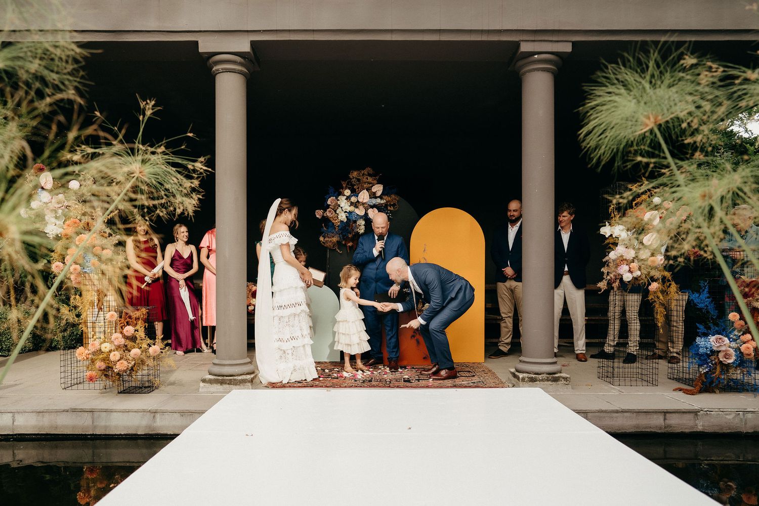 Wedding ceremony held on an elegant porch with decorative pillars and golden floral arrangements at sunset.