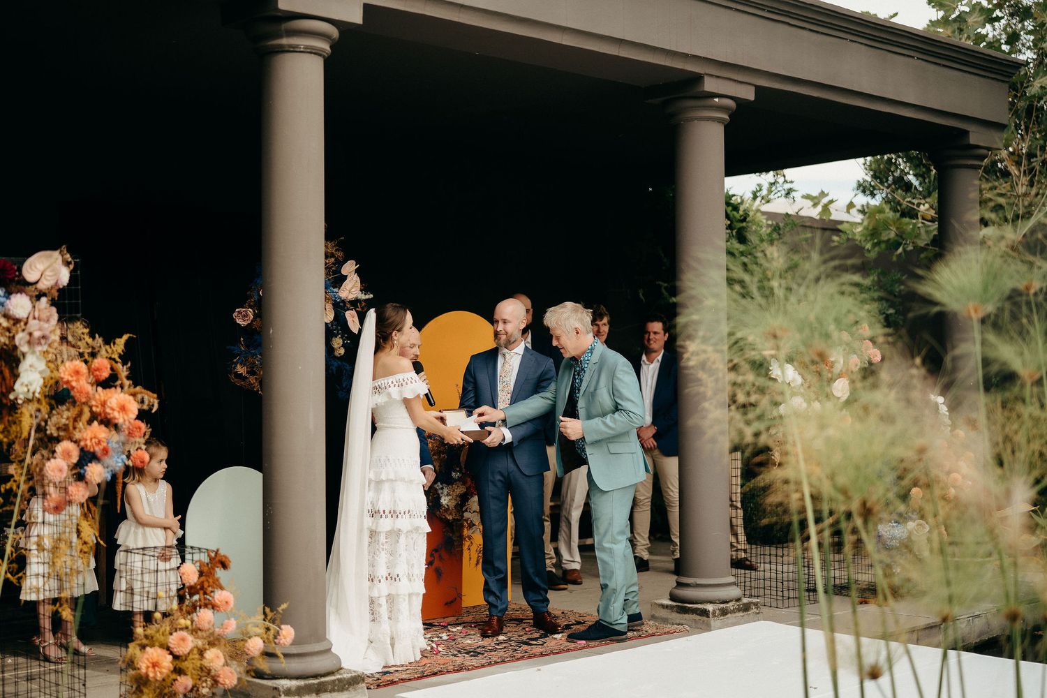 A romantic outdoor wedding ceremony takes place under a columned pavilion decorated with autumn-colored florals.