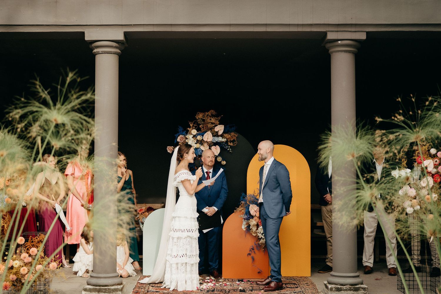 An intimate outdoor wedding ceremony takes place under a columned portico decorated with tropical greenery arrangements.