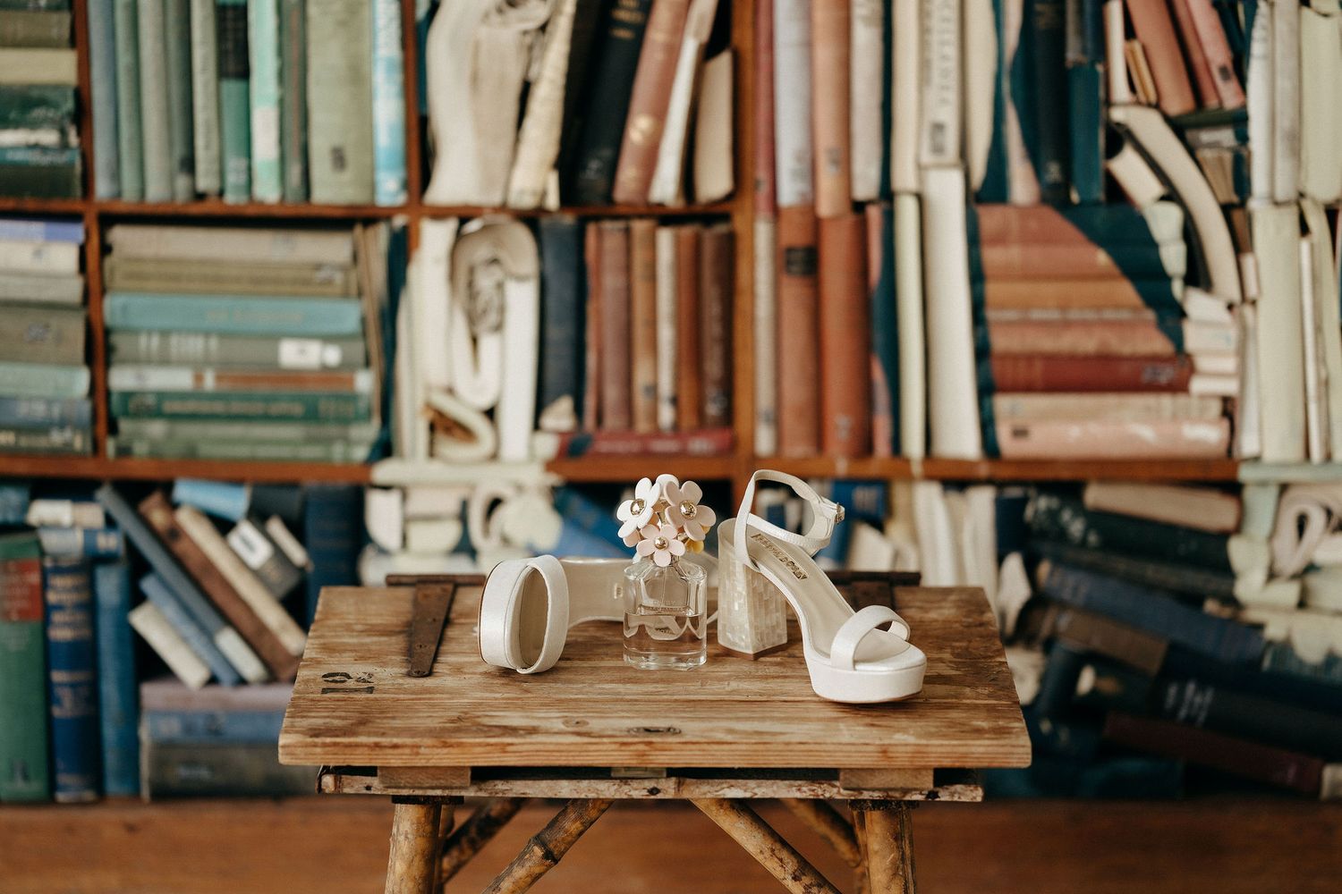 White bridal wedding shoes and jewelry placed on rustic wooden table with blurred bookshelves in background.