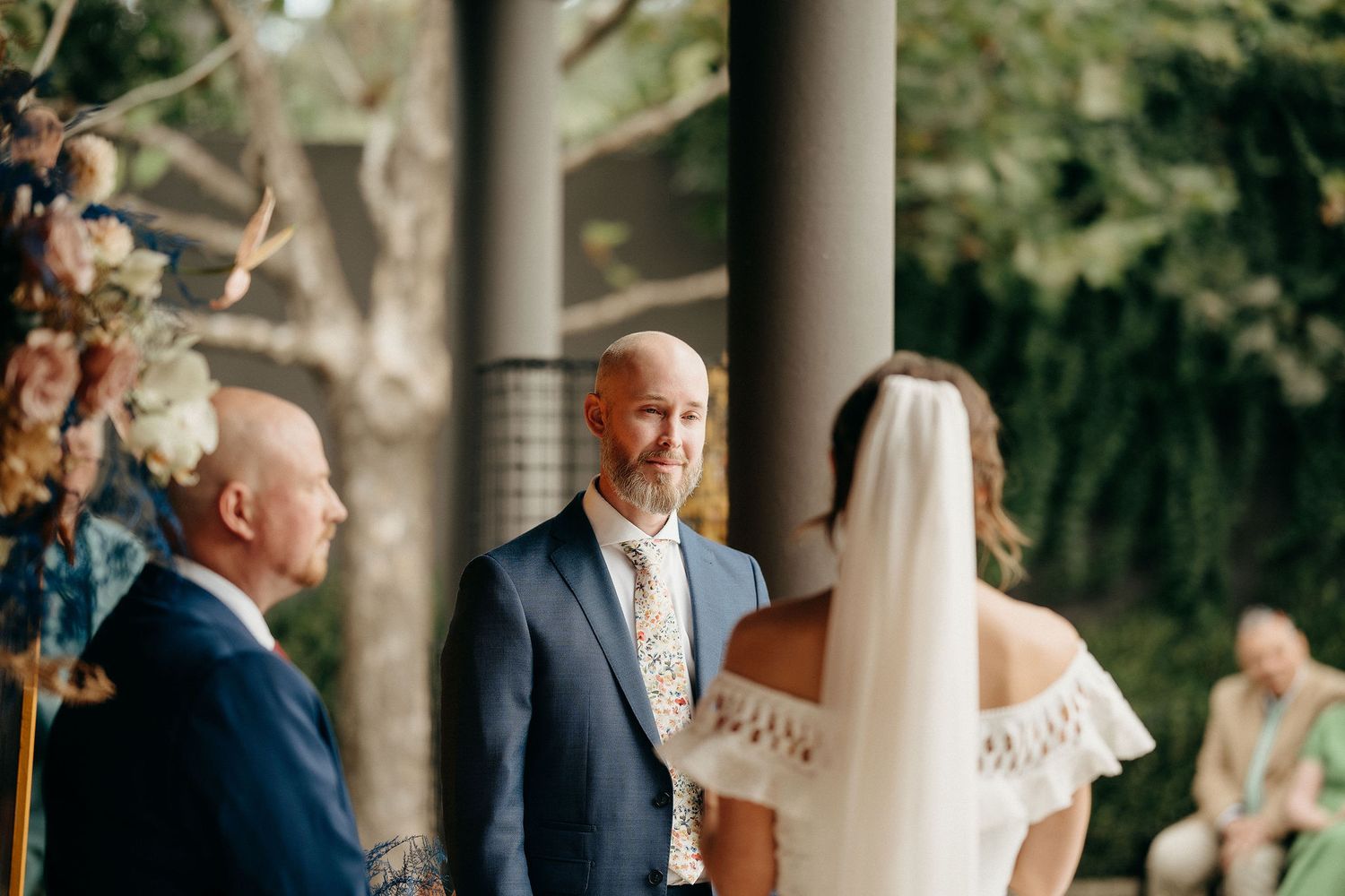 A couple exchanges vows during an outdoor wedding ceremony beneath pillars decorated with floral arrangements.