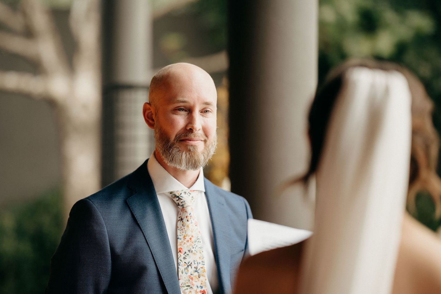Man in navy suit and floral tie smiles warmly during wedding ceremony at columned venue.