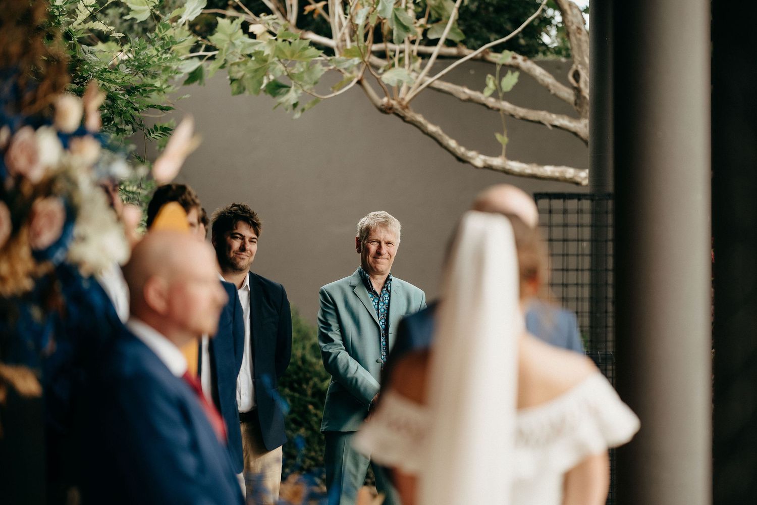 A wedding ceremony outdoors with guests gathered under greenery and branches on a sunny afternoon.