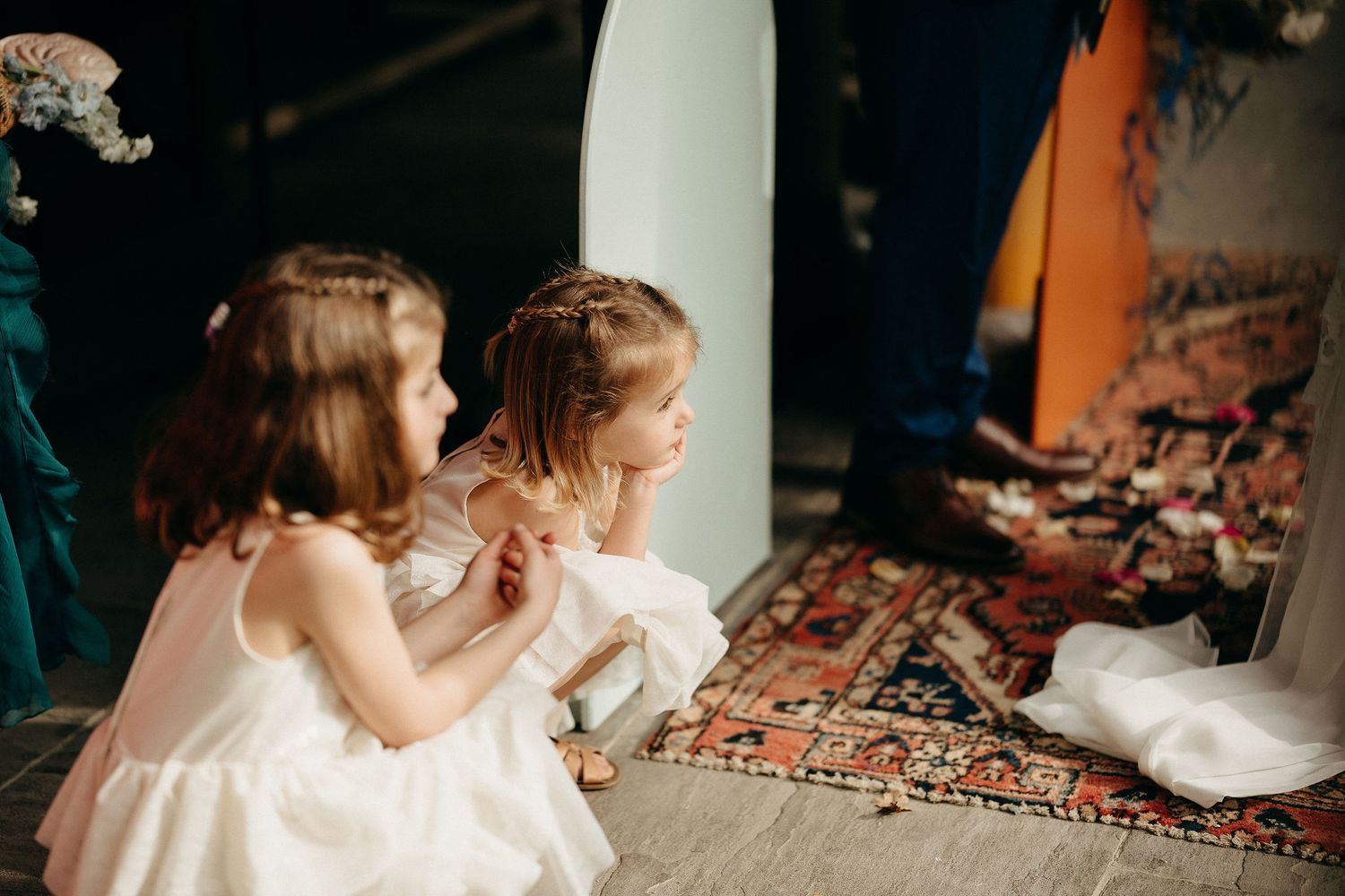 Two small figures in white dresses sit together on a patterned carpet in a dimly lit room.