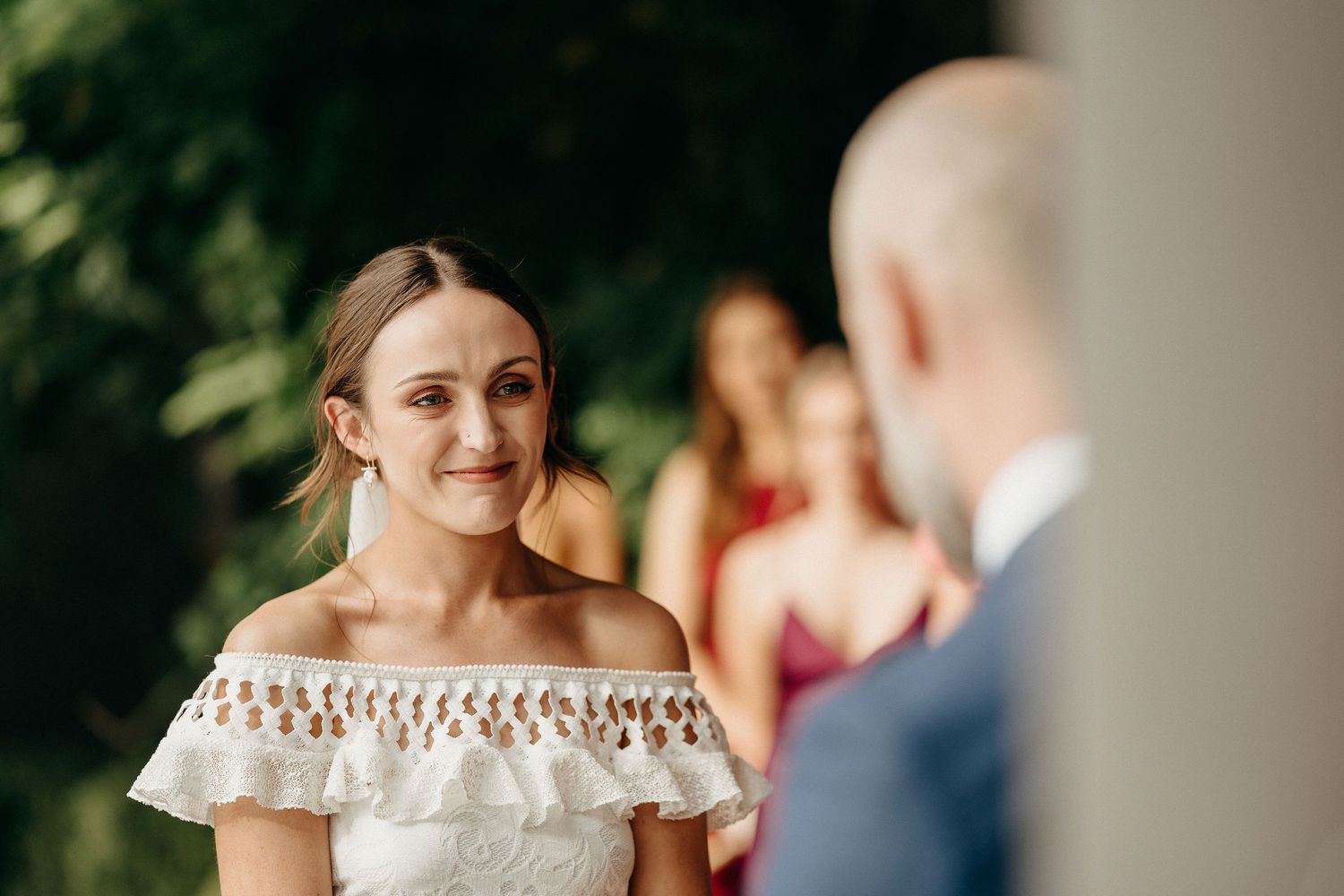 A bride in an off-the-shoulder ruffled wedding dress smiles warmly during an outdoor ceremony.