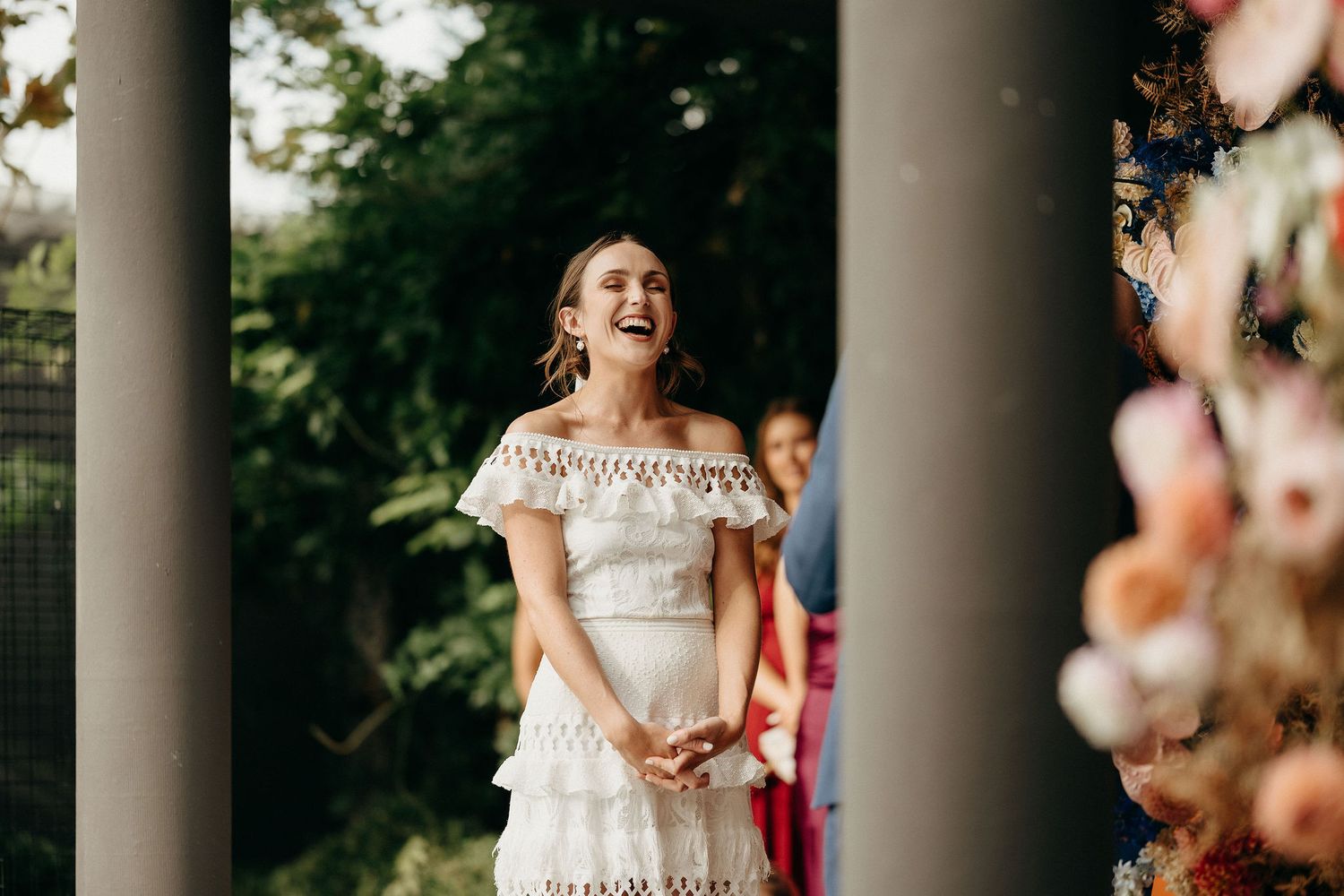 A joyful bride wearing an off-shoulder lace dress laughs while standing between elegant columns at an outdoor venue.