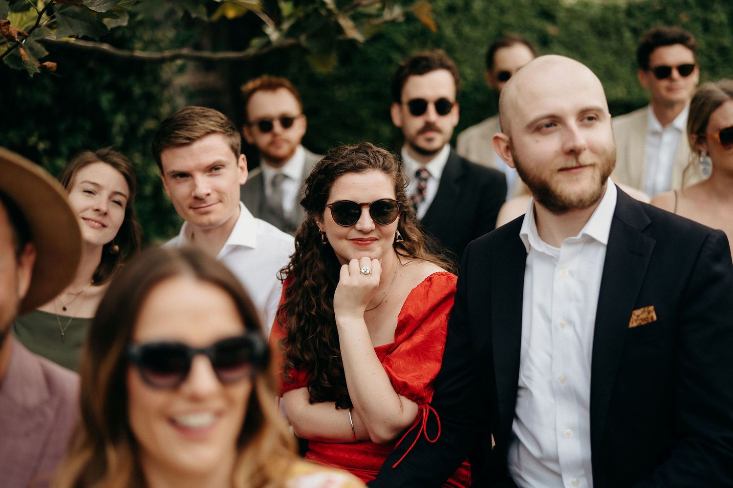 Group of wedding guests gather outdoors, with some wearing sunglasses and one person in a bright red dress.