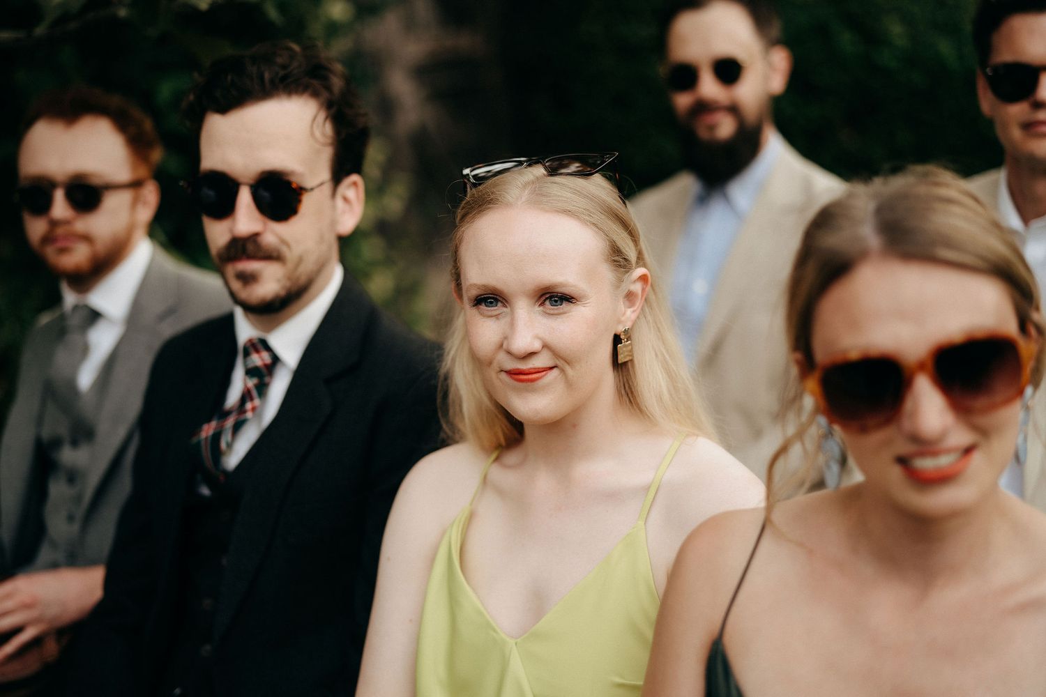Wedding guests in sunglasses gather outdoors on a sunny day, with someone wearing a pale yellow dress in focus.