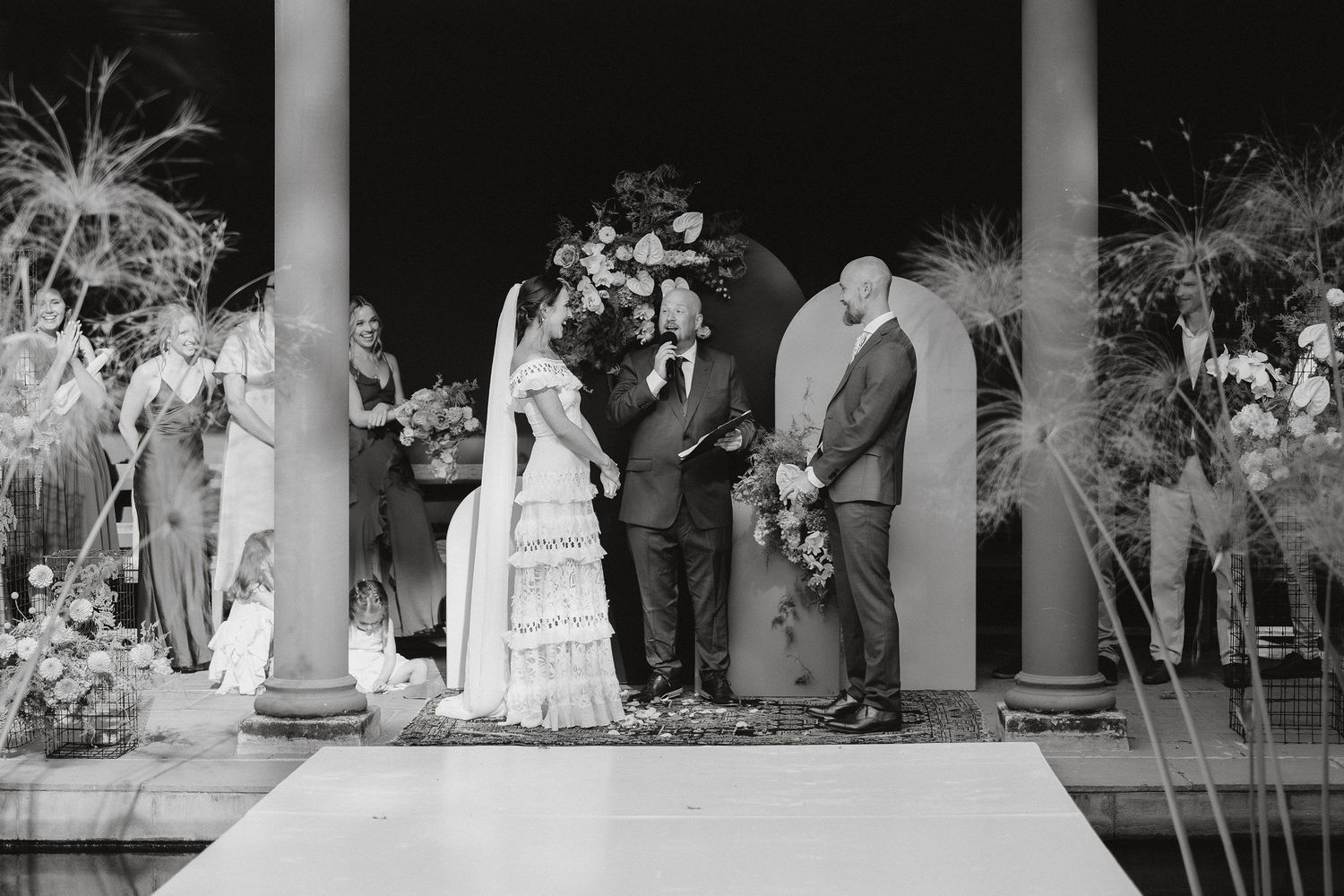 A black and white wedding ceremony taking place under a columned structure with plants and decorations.