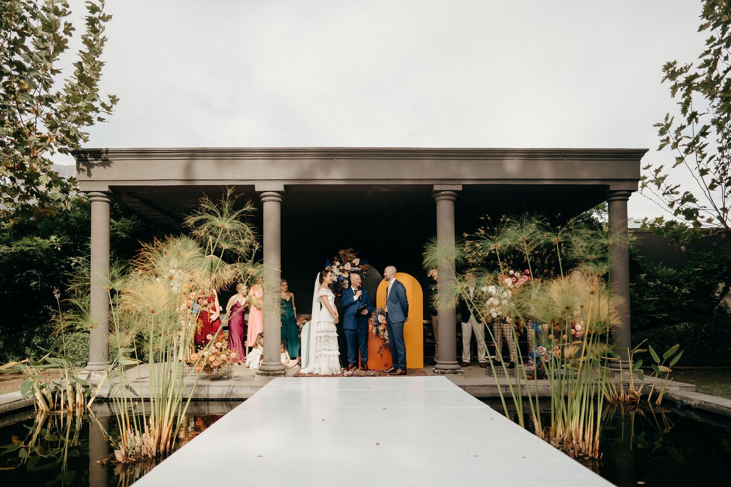 A wedding party stands under an elegant pergola surrounded by lush greenery and ornamental grasses.