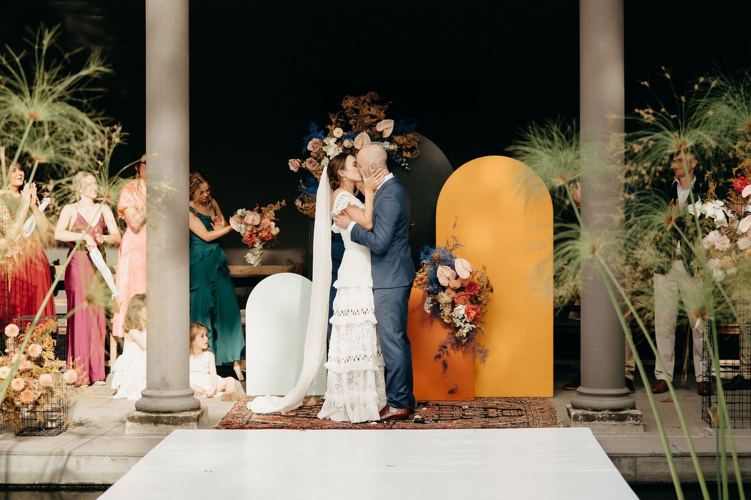 A romantic wedding ceremony takes place under a columned archway with a decorative orange backdrop and tropical palms.
