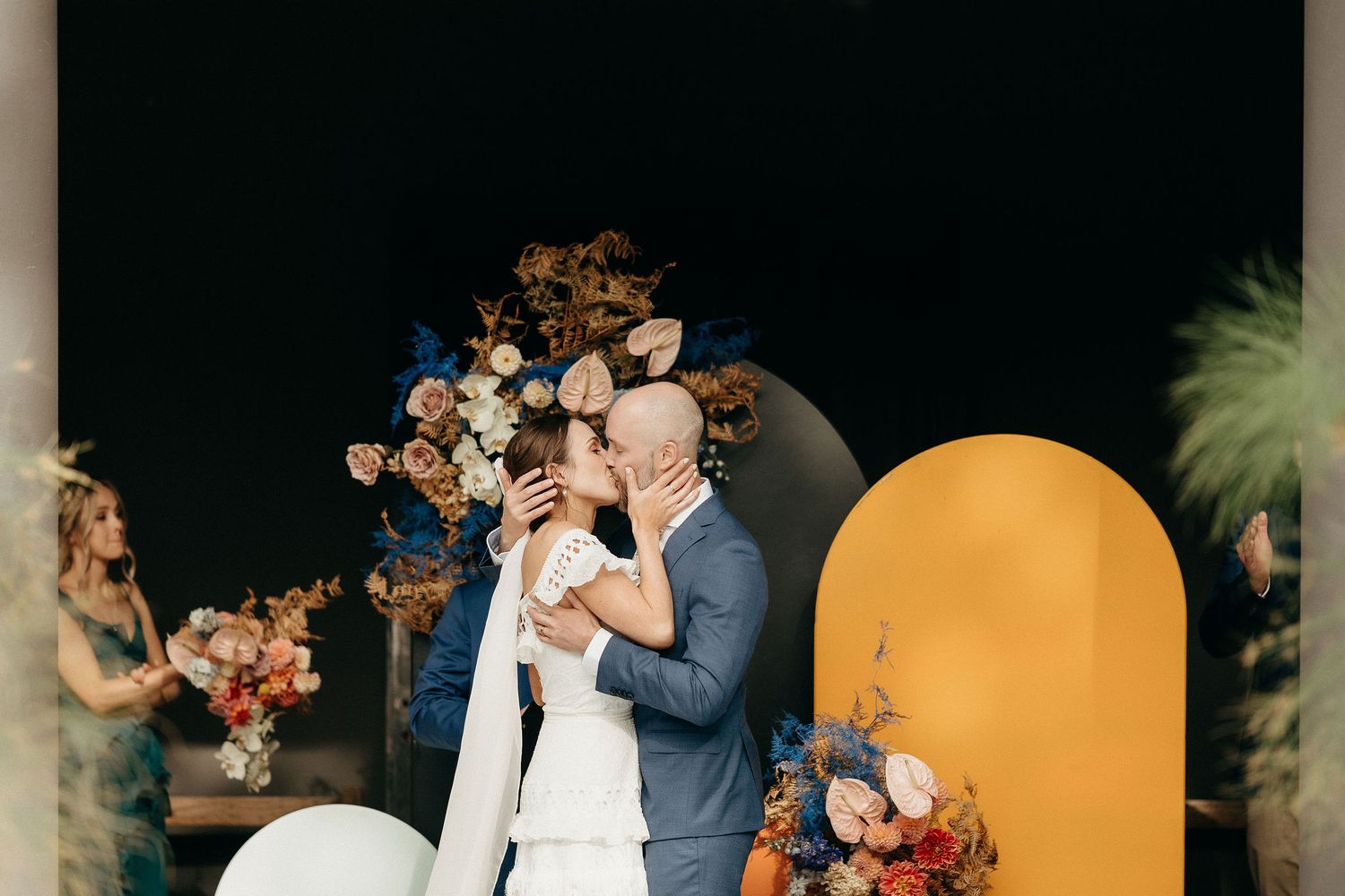 A romantic wedding moment captured at a ceremony arch with orange and navy decor against a dark backdrop.