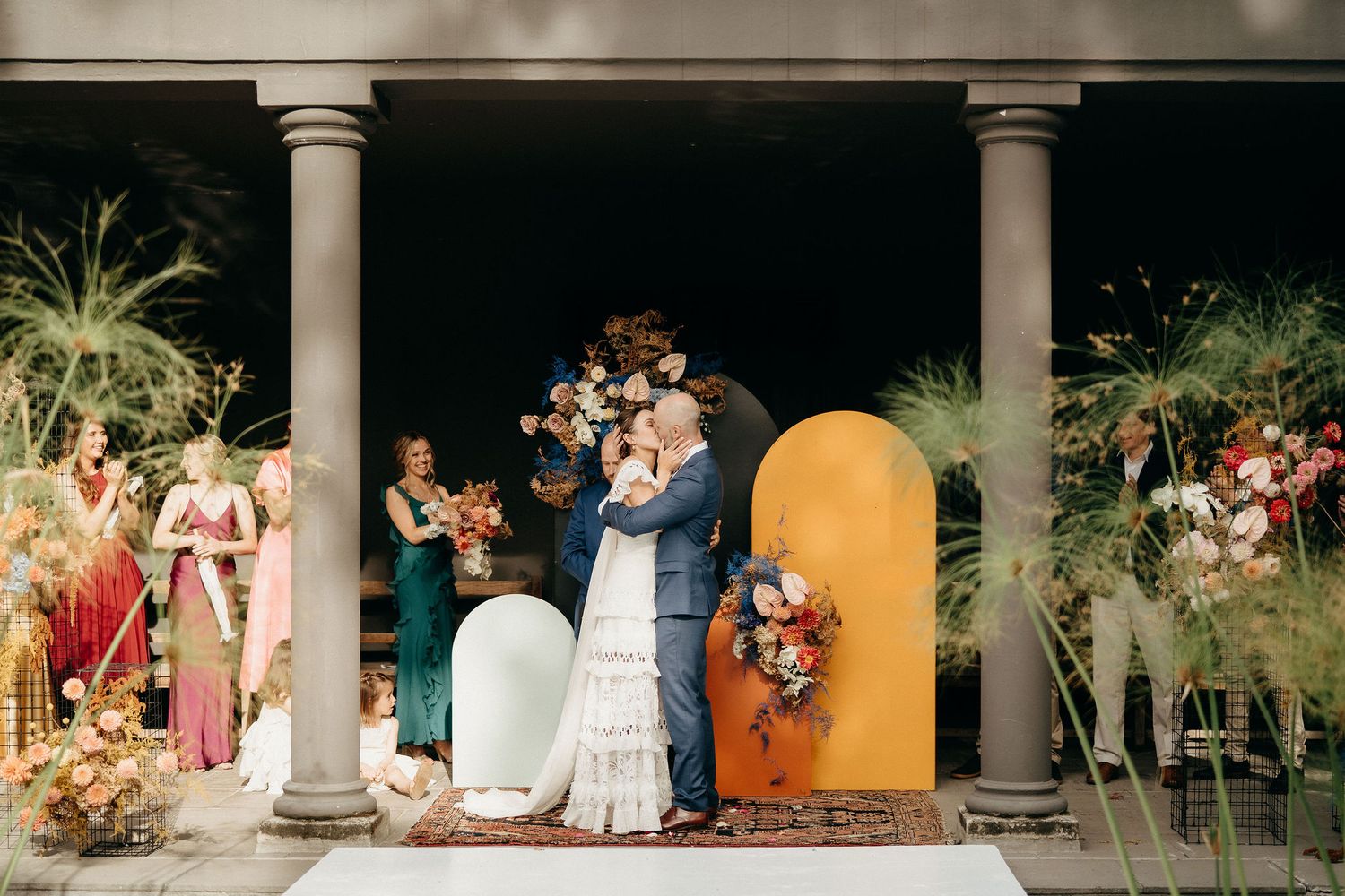 Wedding ceremony under columned archway with colorful arch backdrop and tropical greenery decor.