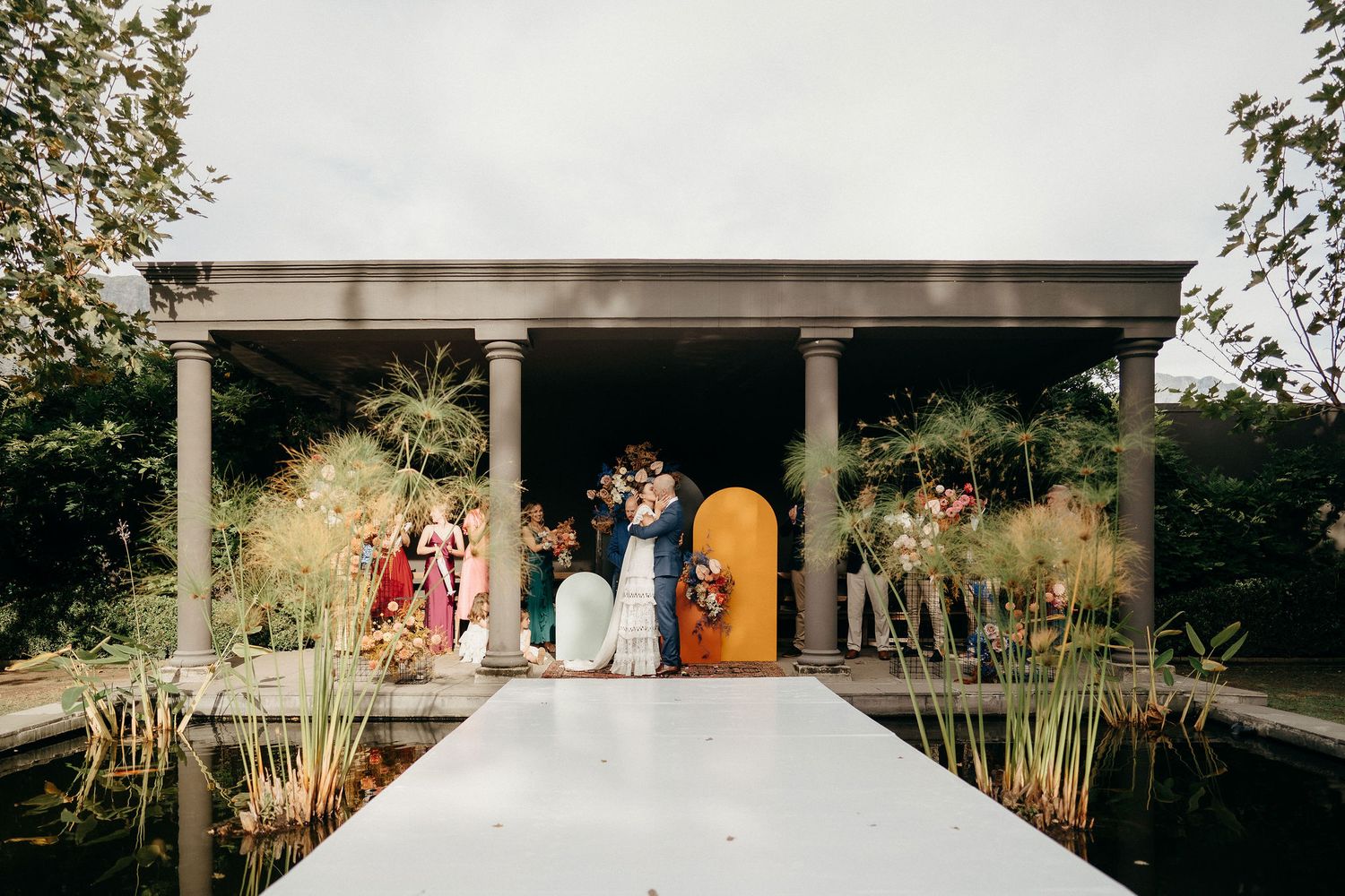 Wedding party stands under an elegant outdoor pavilion with pillars surrounded by lush greenery and a white aisle runner.
