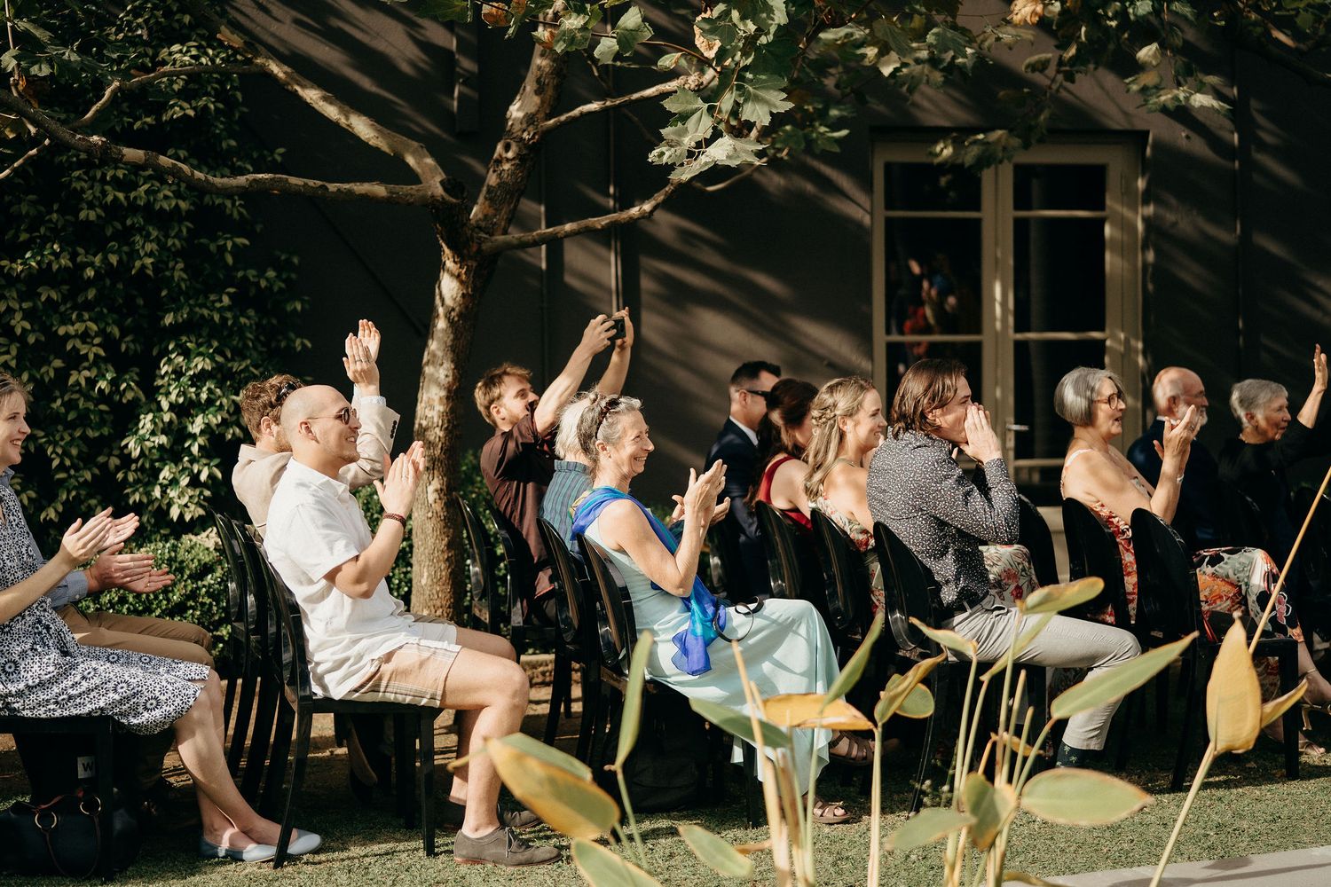 Wedding guests sit in chairs on a lawn during an outdoor celebration as they react joyfully to something offscreen.