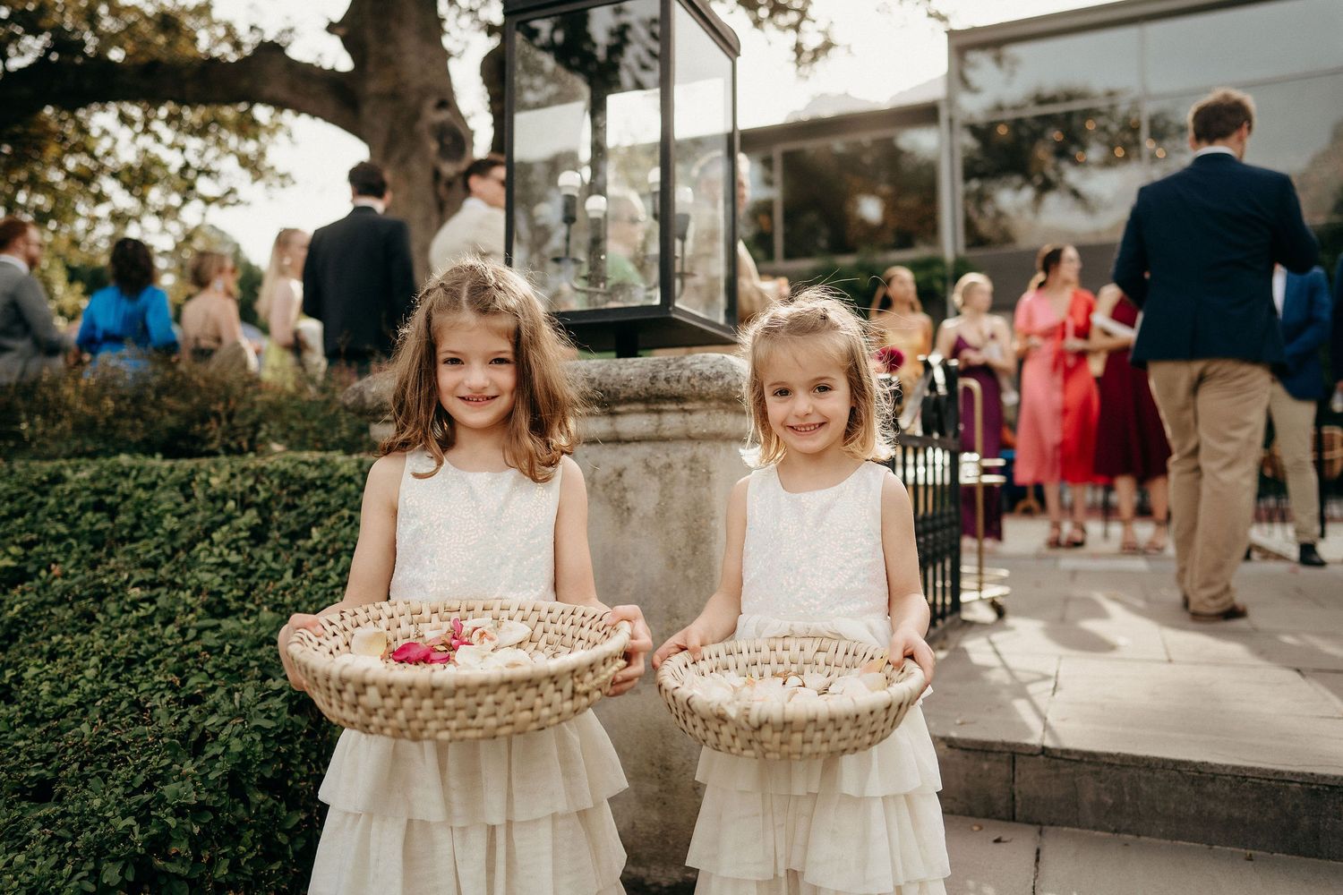 Two flower girls at a wedding holding woven baskets filled with petals while standing together in matching white dresses.