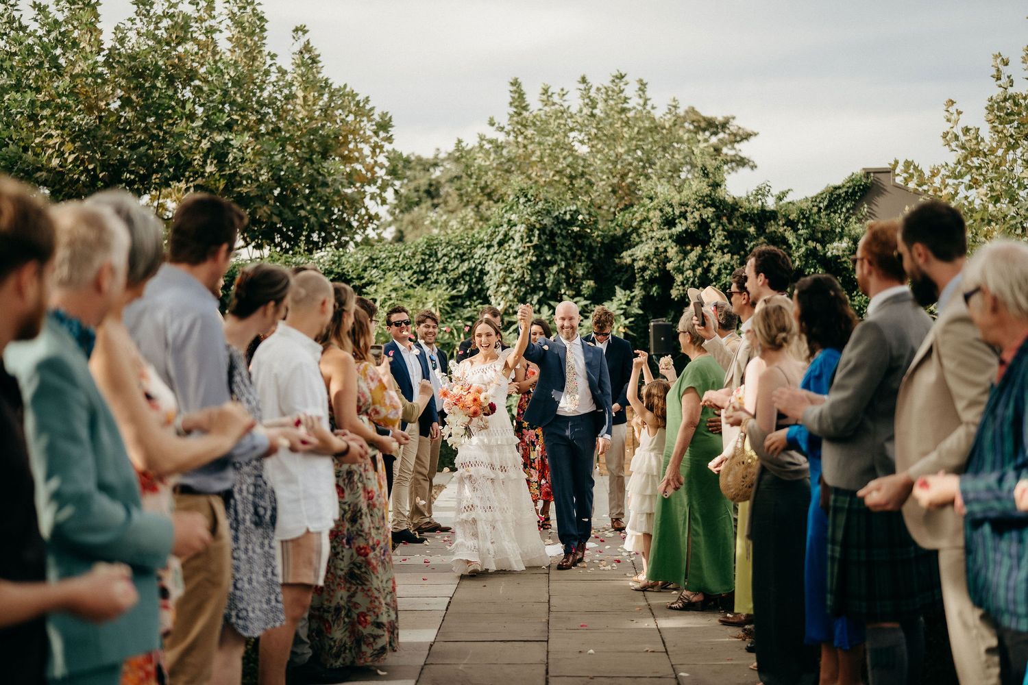 Wedding guests line a garden path as newlyweds walk through during an outdoor ceremony recessional.