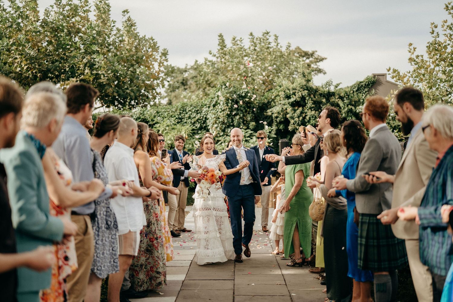 Wedding guests line a garden pathway as newlyweds walk between them during an outdoor summer celebration.