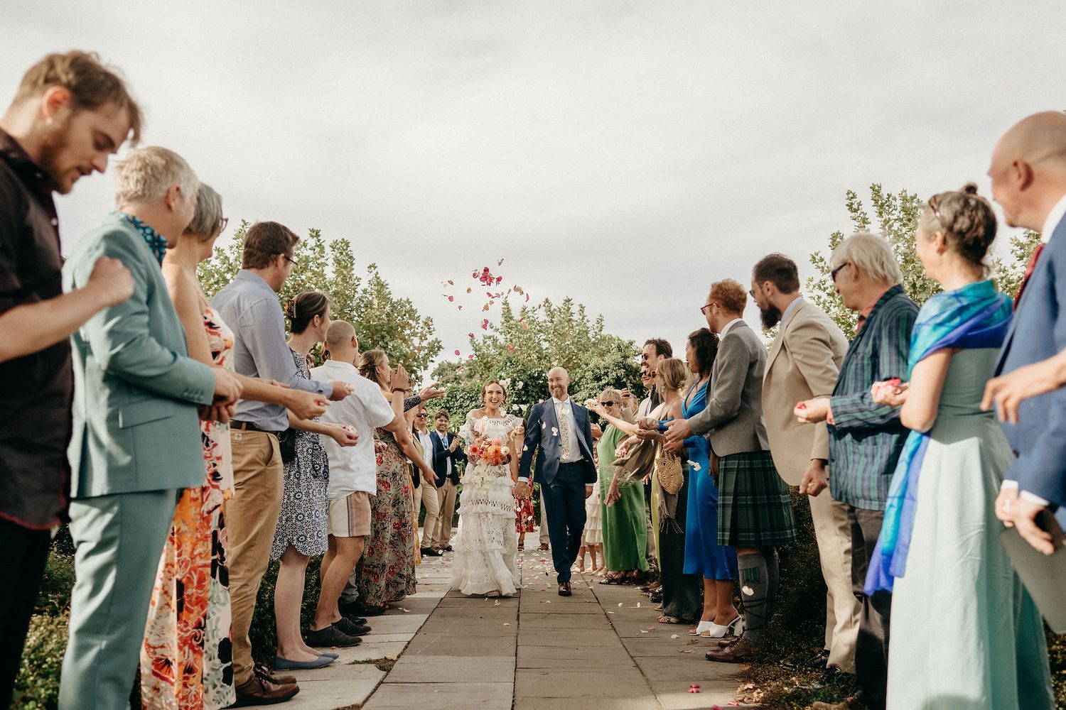 Wedding guests form two rows on pathway as flower petals are tossed during an outdoor ceremony celebration.