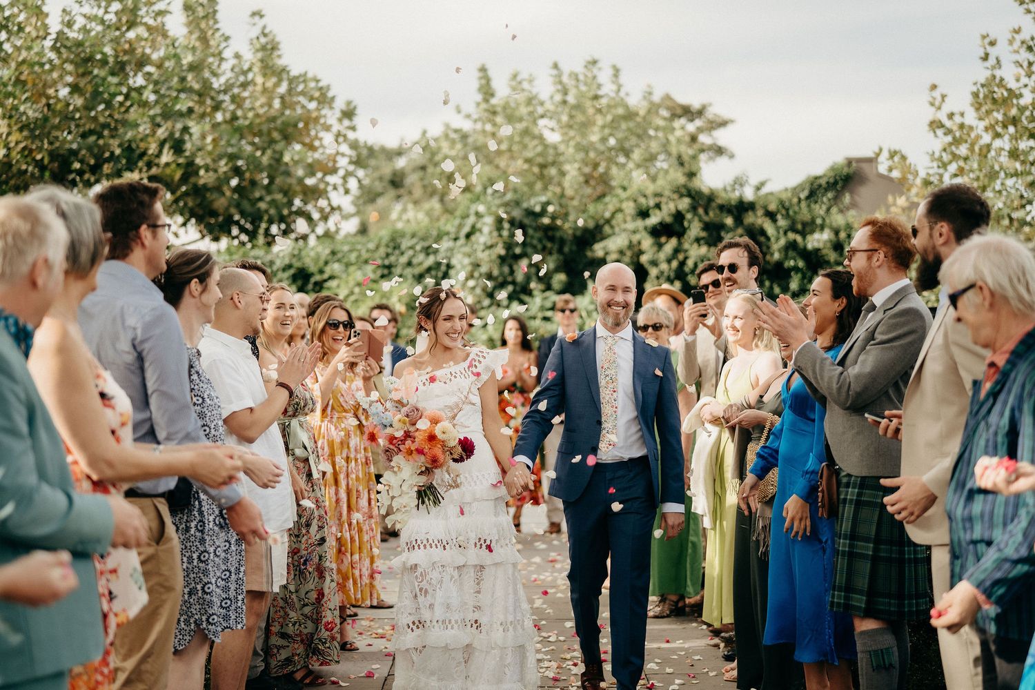 Outdoor wedding ceremony guests stand in two rows during a ceremony at a garden venue on a sunny day.