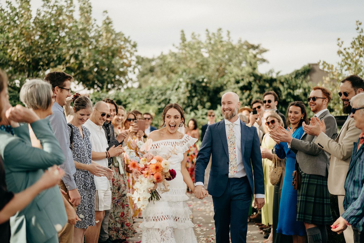 Wedding celebration with guests throwing flower petals at a couple walking down an outdoor path in natural sunlight.