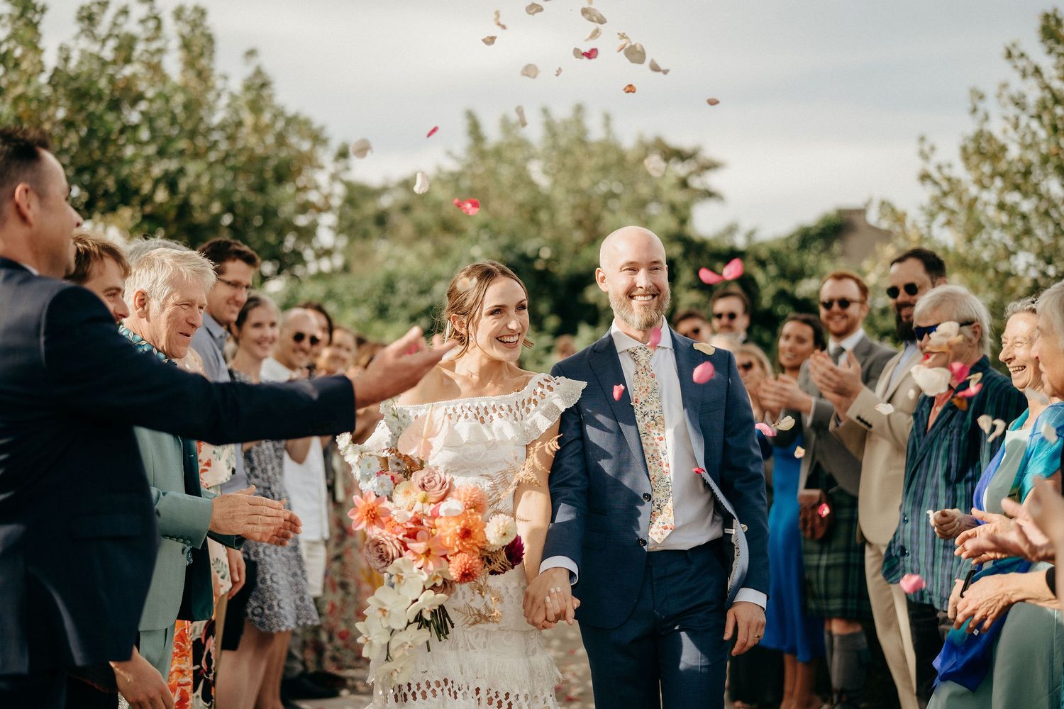 Two newlyweds walk down an aisle lined with guests throwing confetti while celebrating at an outdoor wedding celebration.