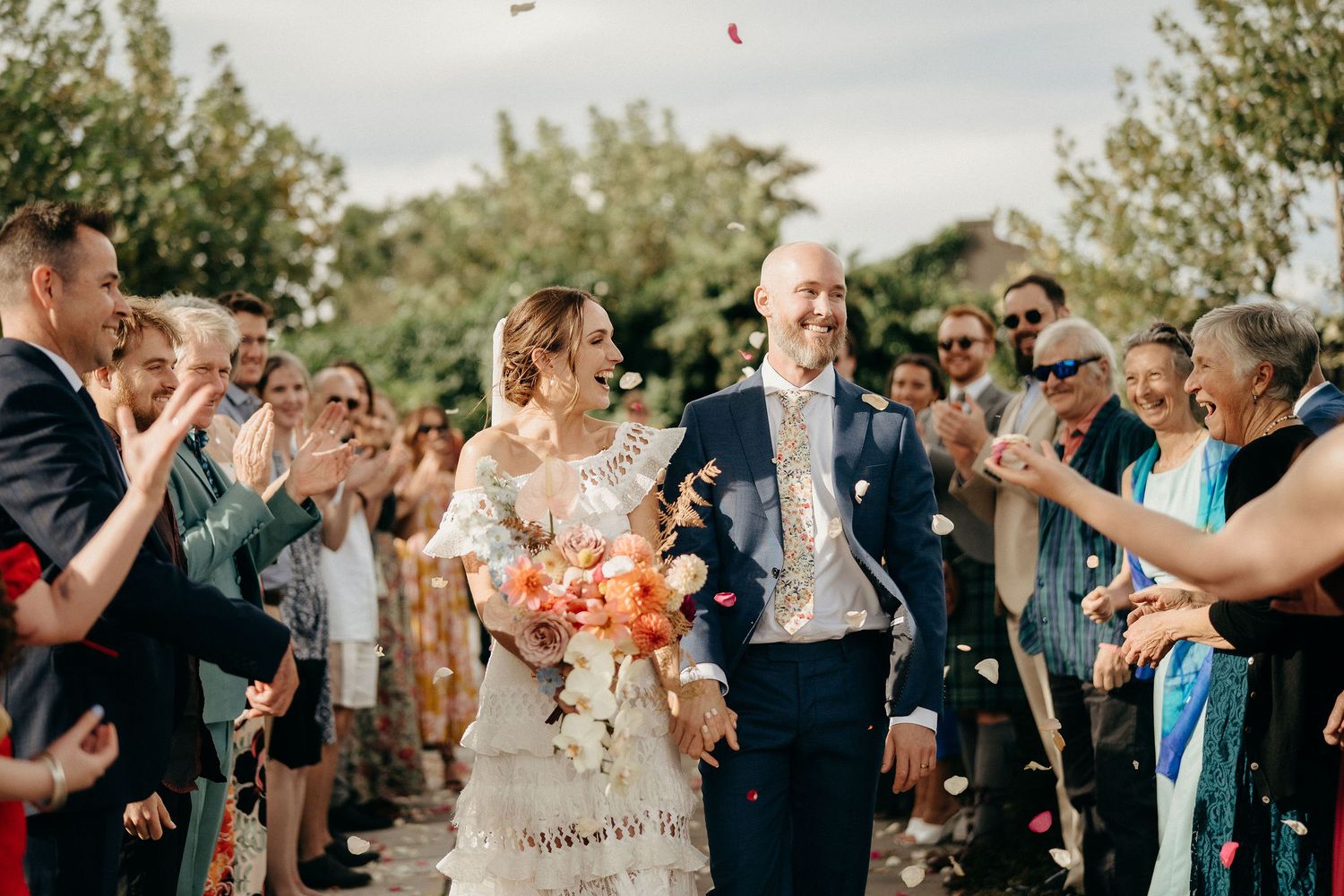 Wedding guests throw petals and celebrate as newlyweds exit down a pathway during a joyful outdoor celebration.