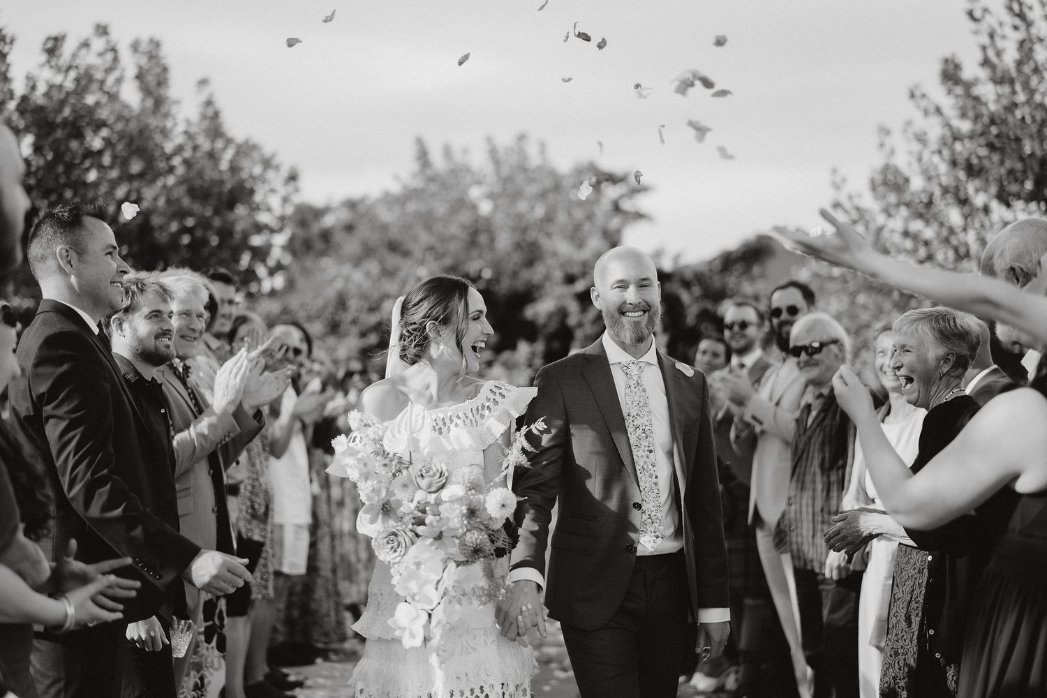 A joyful black and white photo of wedding guests showering the newlyweds with confetti during an outdoor celebration.