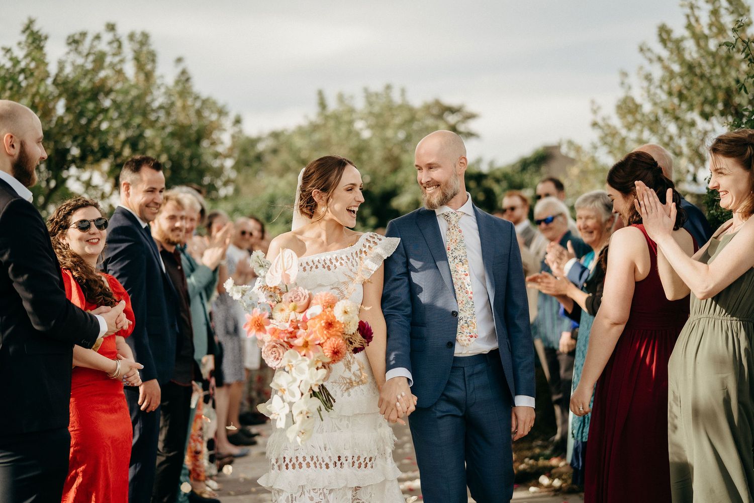Wedding guests line a pathway outdoors while celebrating with flower petals and cheers during a joyful ceremony.