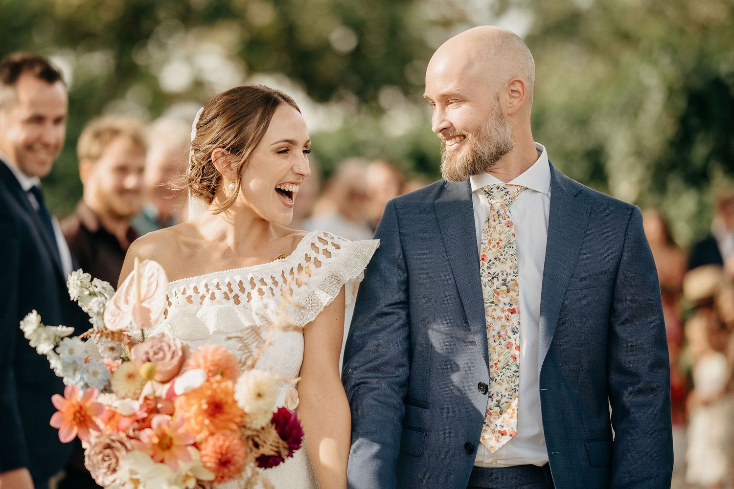 Newlyweds share a joyful moment at an outdoor wedding ceremony with a colorful coral and burgundy bouquet.
