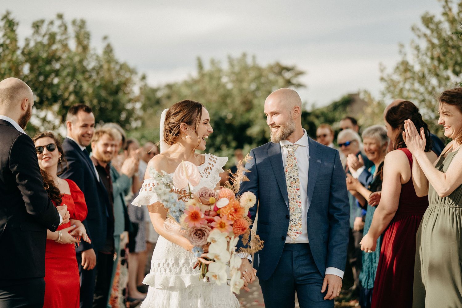 Joyful wedding celebration outdoors with guests looking on as newlyweds share a happy moment holding coral flowers.