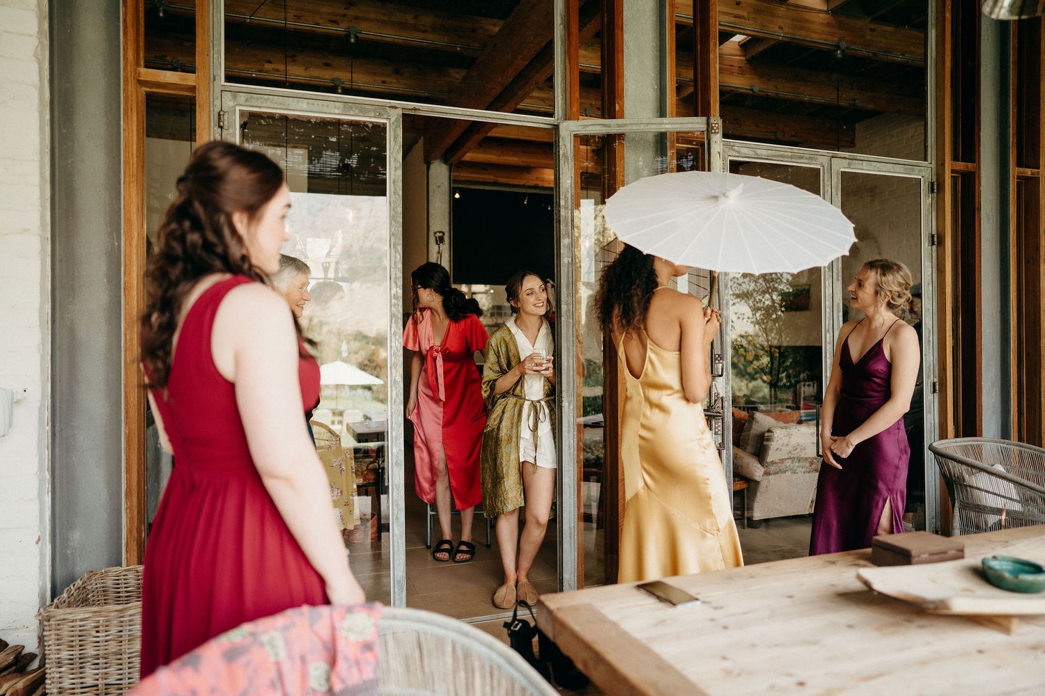 People in formal attire gather on a covered porch, with some holding white parasols during a summer event.