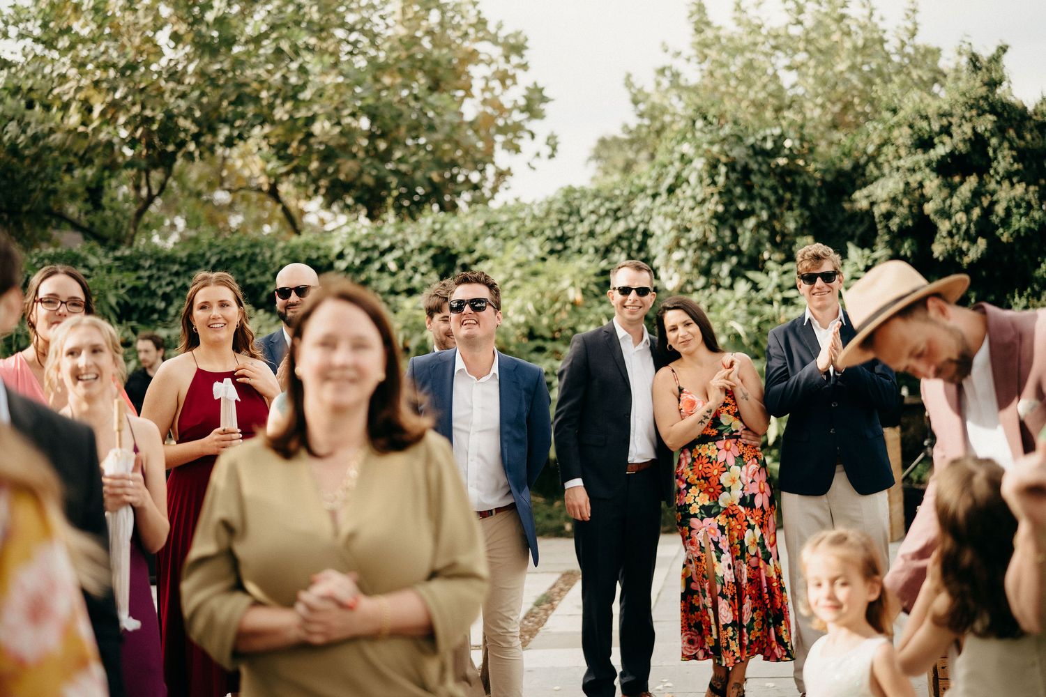 A crowd of wedding guests in summer attire gathering outdoors under shady trees during a daytime celebration.