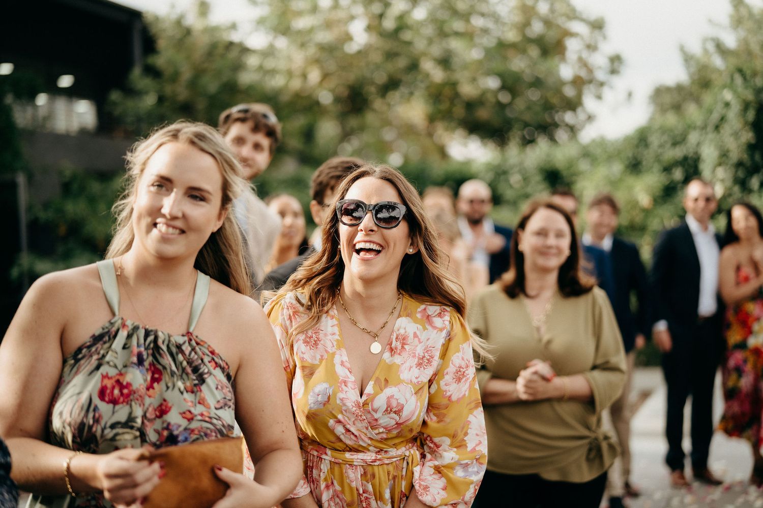 Guests laughing together outdoors at a summer social gathering wearing floral dresses and sunglasses.