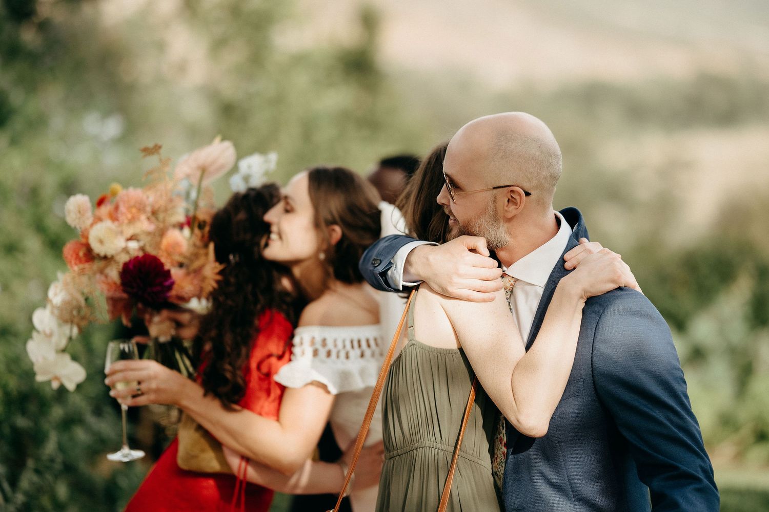 People at an outdoor celebration share laughter and joyful moments while holding a beautiful bouquet of peach-colored flowers.