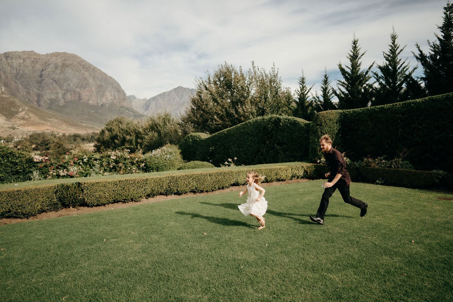 Two children run playfully on a manicured lawn with mountains and evergreen trees in the scenic background.