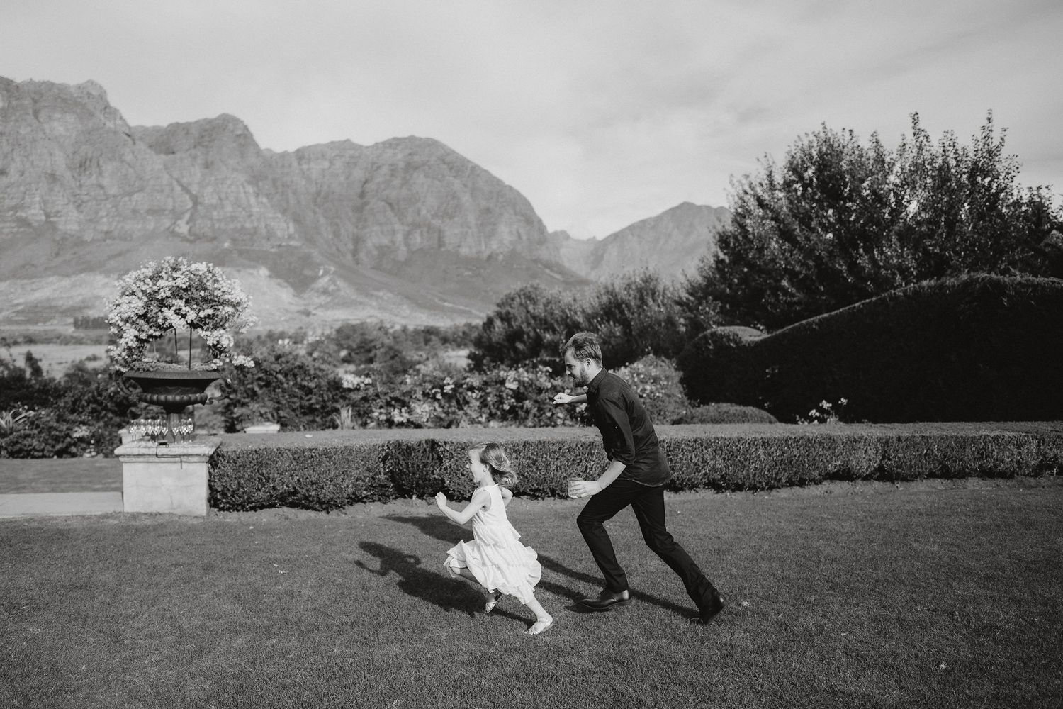 A playful moment captured in black and white as two figures run on a lawn with dramatic mountains in the background.