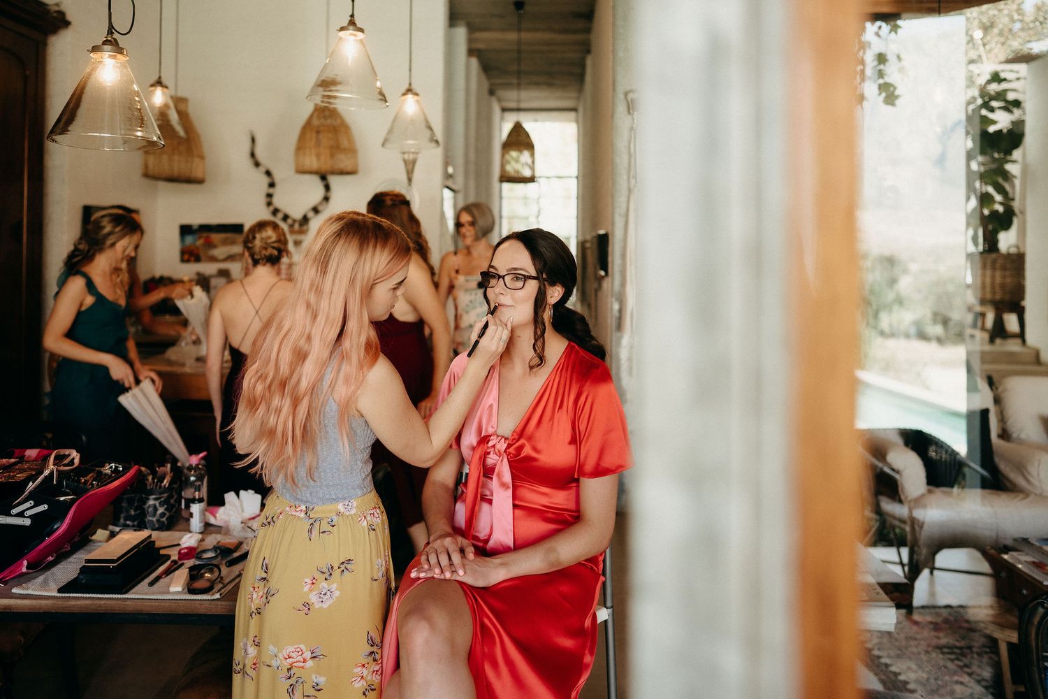 Makeup artist applying cosmetics in a bright salon with hanging pendant lights and warm natural lighting.