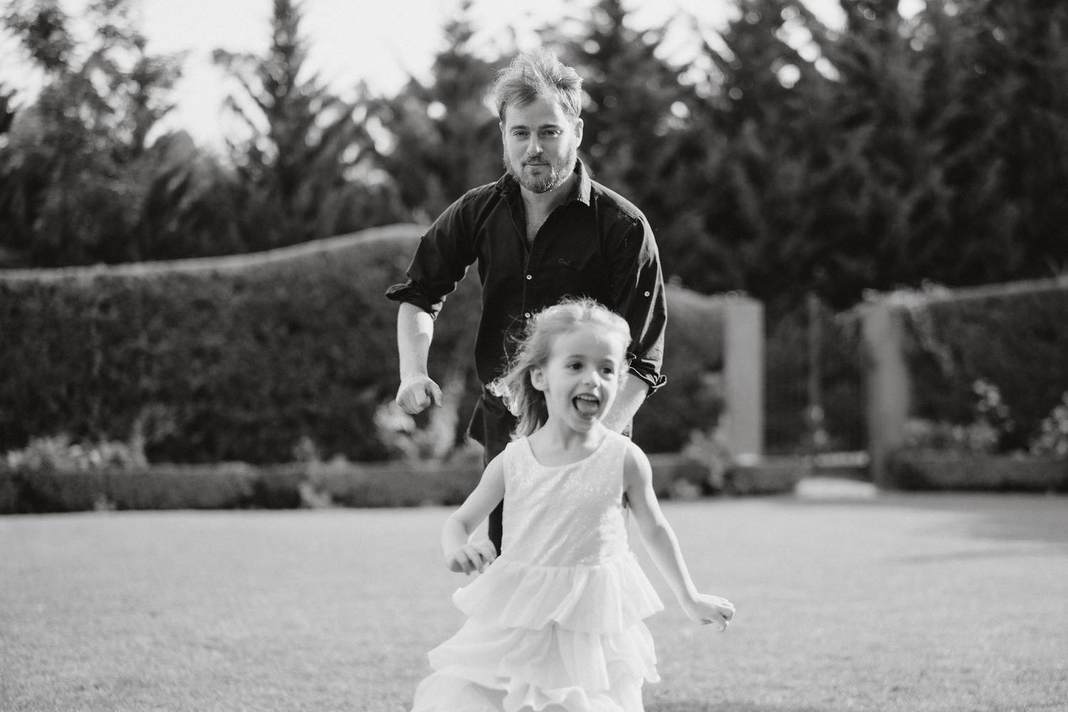 Black and white photo of a child in a white dress running with joy while being chased in a garden setting.