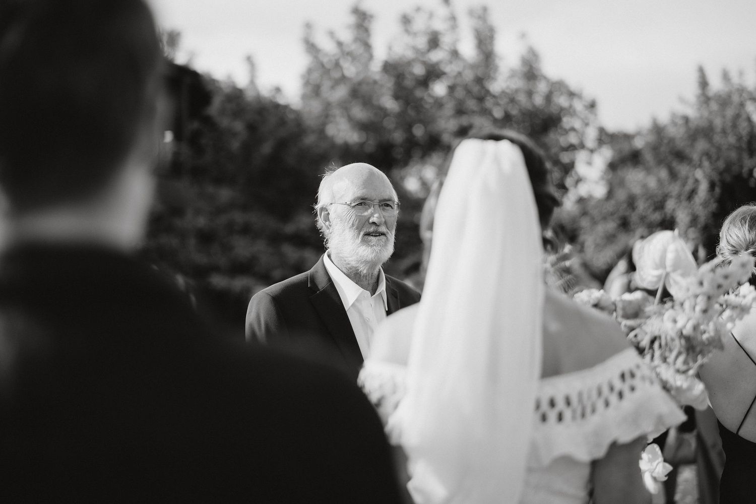 A black and white candid moment captured during an outdoor wedding ceremony with guests gathered in a garden setting.