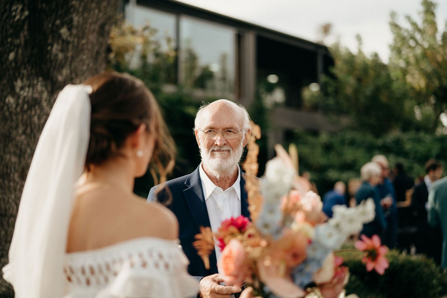 An emotional wedding moment outdoors surrounded by bright floral arrangements in warm sunlight.